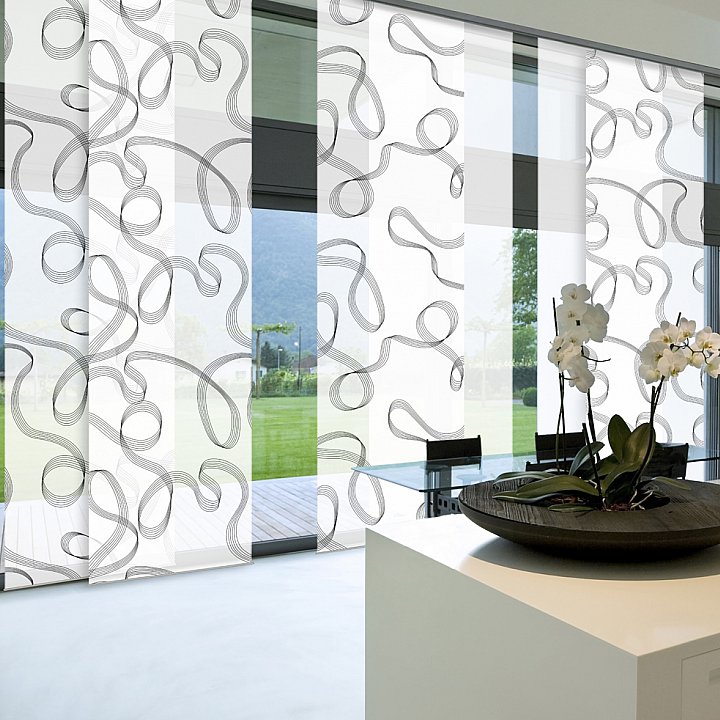 Heller, minimalistischer Raum mit Flächenvorhängen mit einem geschwungenen Muster. Foto von Germania.